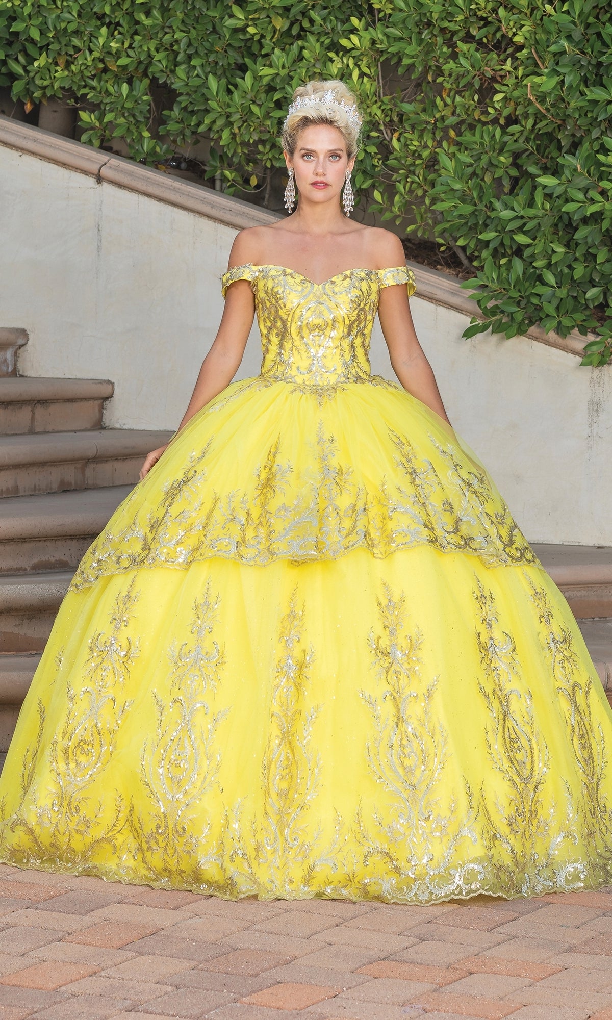 A woman stands on steps in the Quinceanera Dress 1729 By Dancing Queen, a yellow ball gown with gold lace embroidery, off-the-shoulder neckline, crown, and large earrings, greenery behind her.